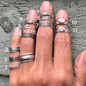 Op de afbeelding: Een collectie van elf zilverkleurige ringen gedragen aan de vingers, genummerd van één tot elf. De ringen hebben diverse ontwerpen, waaronder gladde banden, gedraaide texturen, hartmotieven, dolfijnpatronen, spiraalvormen, bloemmotieven en ovale steenzettingen.