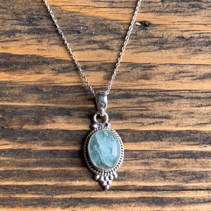 Op de afbeelding: Een zilveren ketting met een lichtblauwe ovale hanger. De hanger is gezet in een zilveren frame met een parelmoerontwerp.