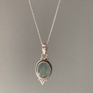 Op de afbeelding: Een zilveren ketting met een ronde, lichtblauwe edelsteen hanger. De hanger heeft een decoratieve rand en een klein, puntig ontwerp aan de onderkant.