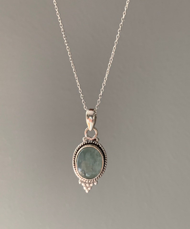 Op de afbeelding: Een zilveren ketting met een ronde, lichtblauwe edelsteen hanger. De hanger heeft een decoratieve rand en een klein, puntig ontwerp aan de onderkant.