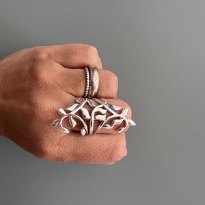 May include: A silver ring featuring a design of interlacing branches and leaves. The ring is worn on a finger.