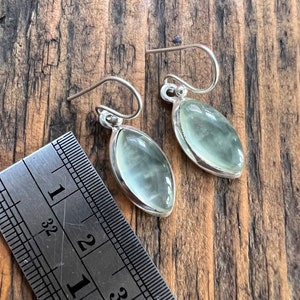 Puede incluir: Un par de pendientes de plata con piedras ovaladas de color verde claro. Los pendientes miden aproximadamente 2,5 cm de largo.