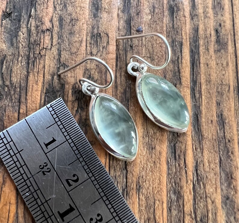 Puede incluir: Un par de pendientes de plata con piedras ovaladas de color verde claro. Los pendientes miden aproximadamente 2,5 cm de largo.