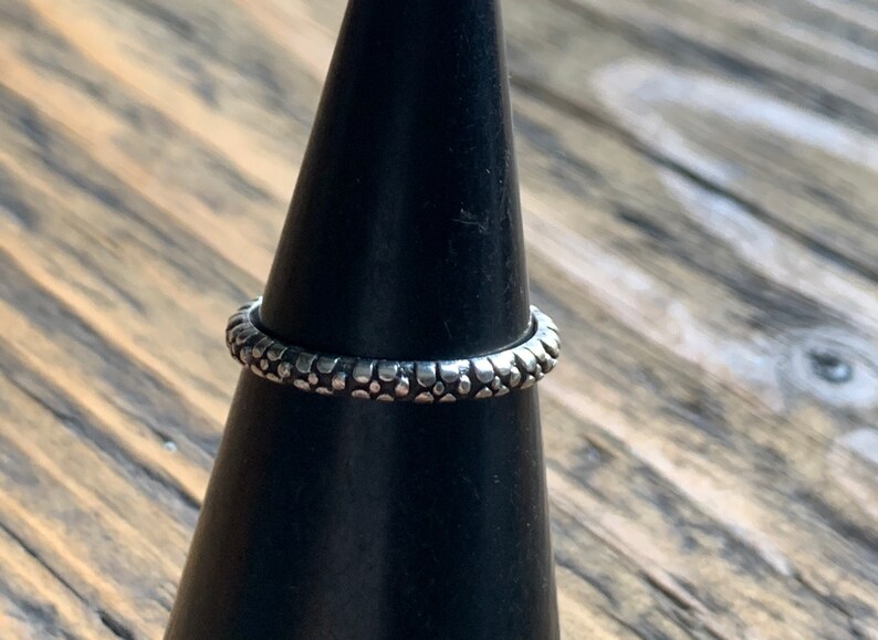 May include: A silver ring with a textured surface. The ring is on a black ring holder.