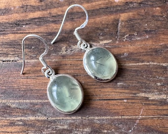 May include: A pair of silver earrings with oval-shaped, pale green gemstones. The gemstones have dark inclusions and are set in a silver bezel. The earrings hang from silver ear wires, displayed on a wooden surface.