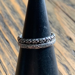 May include: Two silver rings with intricate designs. The top ring has a textured pattern of small dots, while the bottom ring features a swirling, floral design.