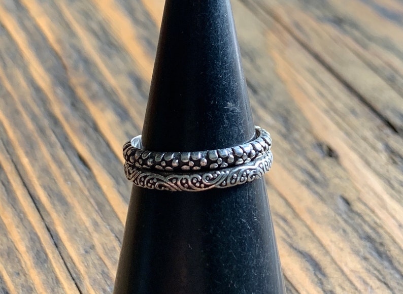 May include: Two silver rings with intricate designs. The top ring has a textured pattern of small dots, while the bottom ring features a swirling, floral design.