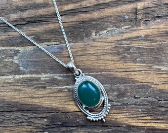 May include: A silver necklace with an oval, dark green gemstone pendant. The pendant is set in a decorative silver frame and hangs from a delicate silver chain. The necklace is displayed on a rustic wooden surface.