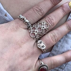 May include: A hand wearing four silver rings. The rings feature a variety of designs, including a chain link, a heart with Chinese characters, a geometric pattern, and a row of elephants. The hand is also wearing a silver chain necklace.