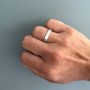 Puede incluir: Un anillo de banda de plata con una forma plana y rectangular.