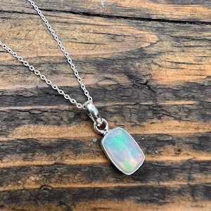 Könnte beinhalten: Eine silberne Halskette mit einem rechteckigen Opalanhänger. Der Opal zeigt schillernde Farben von Grün, Blau und Rosa. Die zarte Kette liegt auf einem Holzgrund. Der Anhänger ist etwa 1,5 cm lang.