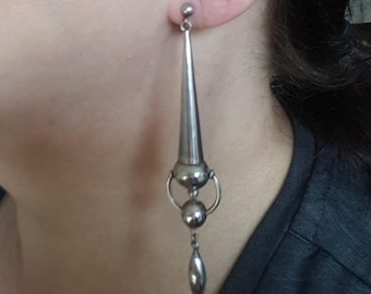May include: A silver-toned earring with a post, a small ball, a ring, two spheres, and a long, pointed, conical pendant. The earring is worn on the ear, and the design is geometric and modern.