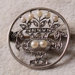 Puede incluir: Un broche de plata con un diseño delicado de un jarrón con flores y perlas. El jarrón está rodeado por un círculo de perlas más pequeñas.