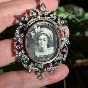 Puede incluir: Un colgante ovalado con un retrato en blanco y negro de una persona con sombrero y cuello de volantes. El marco es de metal plateado adornado con pedrería transparente, rosa y verde en un patrón decorativo de estilo floral.