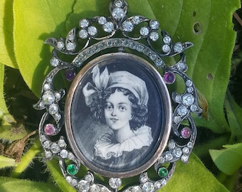 May include: A silver pendant with a black and white portrait of a young person wearing a white hat. The pendant is decorated with small diamonds and green and pink gemstones.