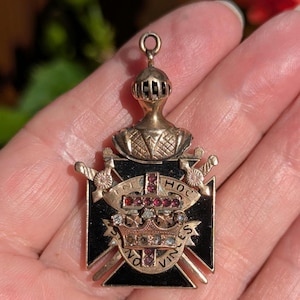 May include: A gold-tone metal pendant featuring a knight's helmet above a black cross emblem. The design includes two crossed swords, a central crown, and a cross set with small red stones. A banner wraps around the center with the text IN HOC VINCES.
