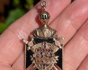 Könnte beinhalten: Ein goldfarbener Metallanhänger mit einem Ritterhelm über einem schwarzen Kreuzemblem. Das Design umfasst zwei gekreuzte Schwerter, eine zentrale Krone und ein mit kleinen roten Steinen besetztes Kreuz. Ein Banner trägt die Inschrift IN HOC VINCES.