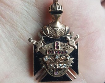 May include: Gold pendant featuring a knight in armor with a black enamel shield. The shield has a cross with red jewels, a crown with clear jewels, and the text "IN HOC SIGNO VINCES".