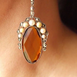 Peut inclure: Une paire de boucles d'oreilles pendantes avec une grande pierre précieuse de couleur ambre entourée d'un halo de petites perles et de pierres de marcassite.
