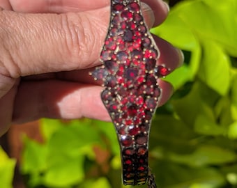 May include: A hinged bangle bracelet featuring a wide, central decorative panel densely set with numerous small, deep red faceted stones. The metal frame is dark in tone, and a thin safety chain is attached to the side of the bracelet.