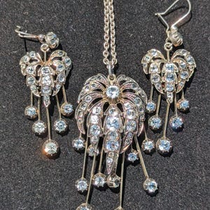 Op de afbeelding: Een zilverkleurige sieradenset bestaande uit een ketting met hanger en twee bijpassende oorbellen. Elk sieraad heeft een waaiervormig ontwerp ingelegd met heldere, sprankelende kristallen en meerdere bungelende kristallen accenten aan dunne kettinkjes.