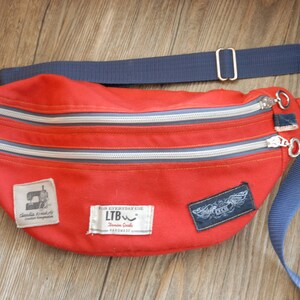 Könnte beinhalten: Rote Canvas-Bauchtasche mit zwei Reißverschlussfächern und einem verstellbaren blauen Gurt. Die Tasche hat drei Patches mit dem Text "Handmade & Crafted", "FOR EVERYDAY USE LTB Dream Goods" und "Angel Wings".