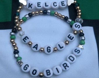 May include: Three gold beaded bracelets with white letter beads spelling out "GO BIRDS", "EAGLES", and "KELCE". The bracelets have green and silver beads as well.