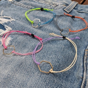 Puede incluir: Seis pulseras de cuerda de colores con un dije hexagonal dorado. Las pulseras se muestran sobre un fondo de mezclilla azul.