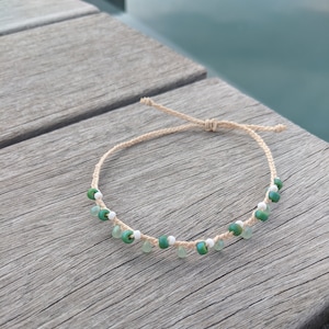 May include: A simple bracelet made with natural twine and green and white beads. The bracelet is tied with a knot and is lying on a wooden surface.