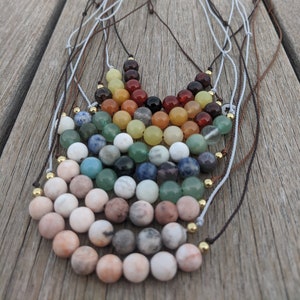 May include: A rainbow of gemstone beads strung on multiple cords, creating a layered necklace. The beads are arranged in a semi-circle shape, with the largest beads at the bottom and the smallest beads at the top. The beads are a variety of colors, including red, orange, yellow, green, blue, and purple.