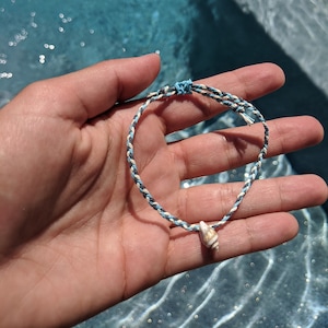 Puede incluir: Un brazalete trenzado azul, blanco y negro con un pequeño dije de concha marina. El brazalete se muestra en una mano contra un fondo de agua azul.