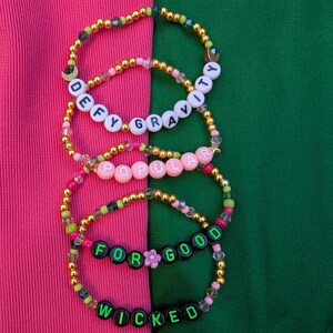 May include: Four beaded bracelets with phrases from the musical "Wicked" and "Defying Gravity". The bracelets are gold, pink, green, and black. The words "Defy Gravity", "Popular", "For Good", and "Wicked" are spelled out with letter beads.