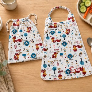 Peut inclure: Un sac à cordon blanc et un bavoir assorti avec un motif répétitif de renards, d'arbres et de textes incluant "Save the Earth" et "Love Forest". Le bavoir est brodé du prénom "Evie" en fil rouge sur le côté inférieur droit.