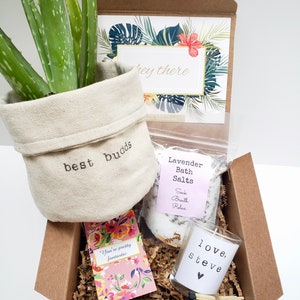 May include: A brown gift box filled with brown shredded paper. The box contains a small potted aloe vera plant in a white canvas pot with the text "best budds" embroidered on it, a small jar of lavender bath salts with the text "Lavender Bath Salts" and "Soak Breath Relax", a small glass candle with the text "love, steve" and a small heart, and a small card with the text "You're pretty fantastic" and floral designs.