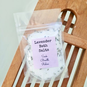 May include: A clear plastic bag filled with white bath salts and lavender buds. The bag has a label that reads "Lavender Bath Salts" and "Soak. Breath. Relax."