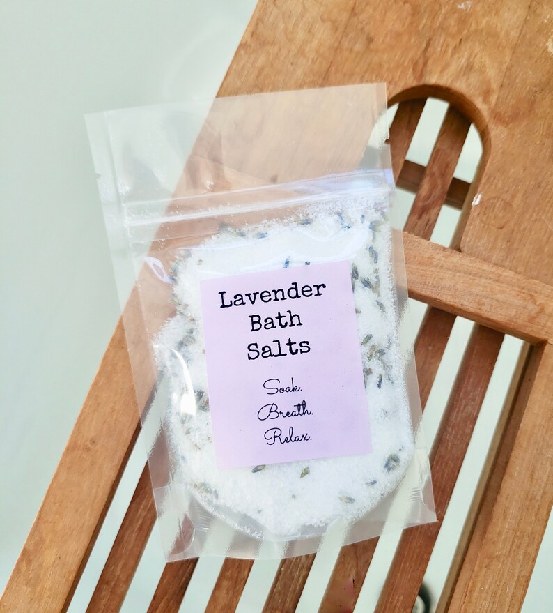 May include: A clear plastic bag filled with white bath salts and lavender buds. The bag has a label that reads "Lavender Bath Salts" and "Soak. Breath. Relax."