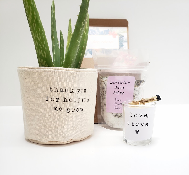 May include: A gift basket with a potted aloe plant, a small candle, and a bag of lavender bath salts. The plant pot has the text "thank you for helping me grow". The candle has the text "love, steve" and a small heart. The bath salts bag has the text "Lavender Bath Salts Sock Breath Relax".
