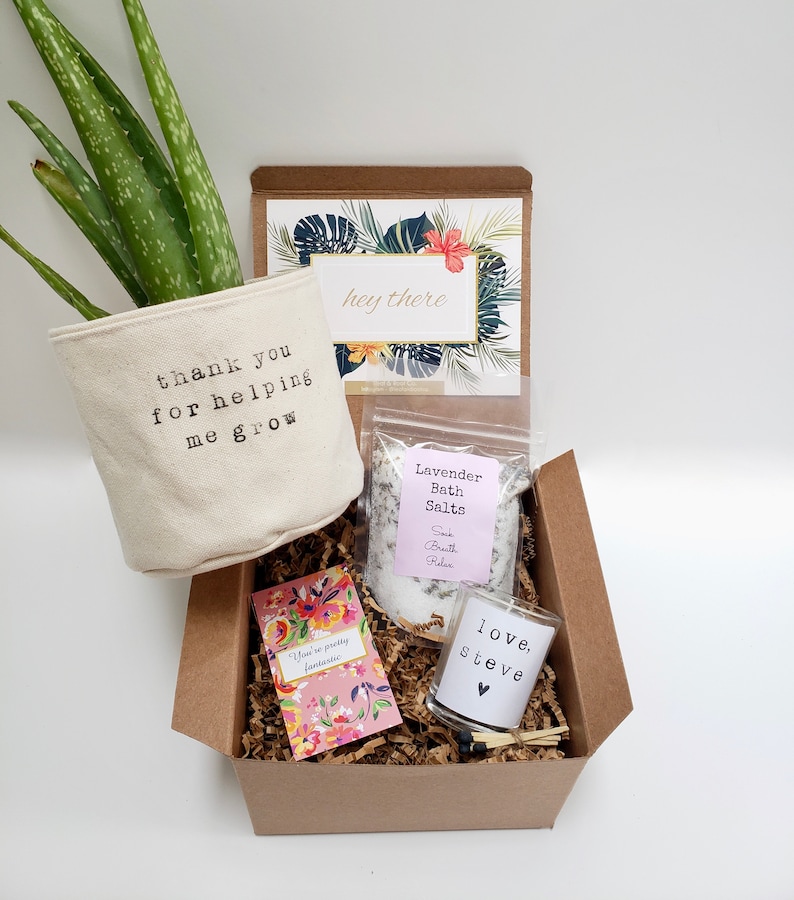 May include: A brown gift box filled with a small potted aloe plant, bath salts, a candle, and a card. The plant pot has the text "thank you for helping me grow". The bath salts are in a clear bag with a pink label that says "Lavender Bath Salts". The candle is in a clear glass jar with a white label that says "love, steve". The card has a floral design and the text "hey there".