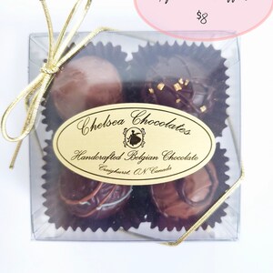 May include: A clear box with a gold ribbon tied around it. Inside the box are four chocolate truffles. The box has a label that says "Chelsea Chocolates" and "Handcrafted Belgian Chocolate".