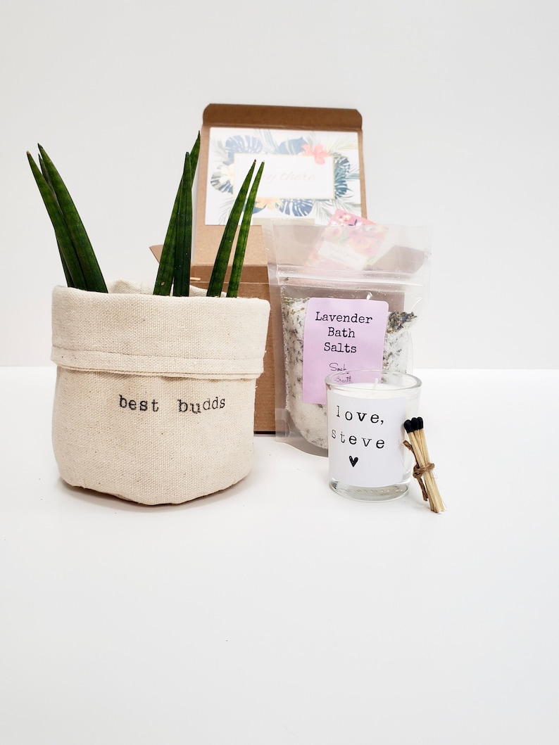 May include: A gift box set with a small potted plant, a bag of lavender bath salts, and a small candle with the text "love, steve" and a heart. The plant pot is made of fabric and has the text "best buds" printed on it.