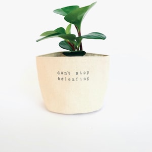Peut inclure: Une petite plante verte dans un pot en tissu blanc avec le texte "don't stop beleafing" imprimé sur le devant.