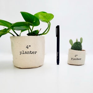 Peut inclure: Deux pots en tissu, l'un de 10 cm de diam&egrave;tre et l'autre de 5 cm de diam&egrave;tre. Le plus grand pot est rempli d'une plante verte, et le plus petit pot est rempli d'un cactus. Les deux pots sont blancs avec du texte noir.