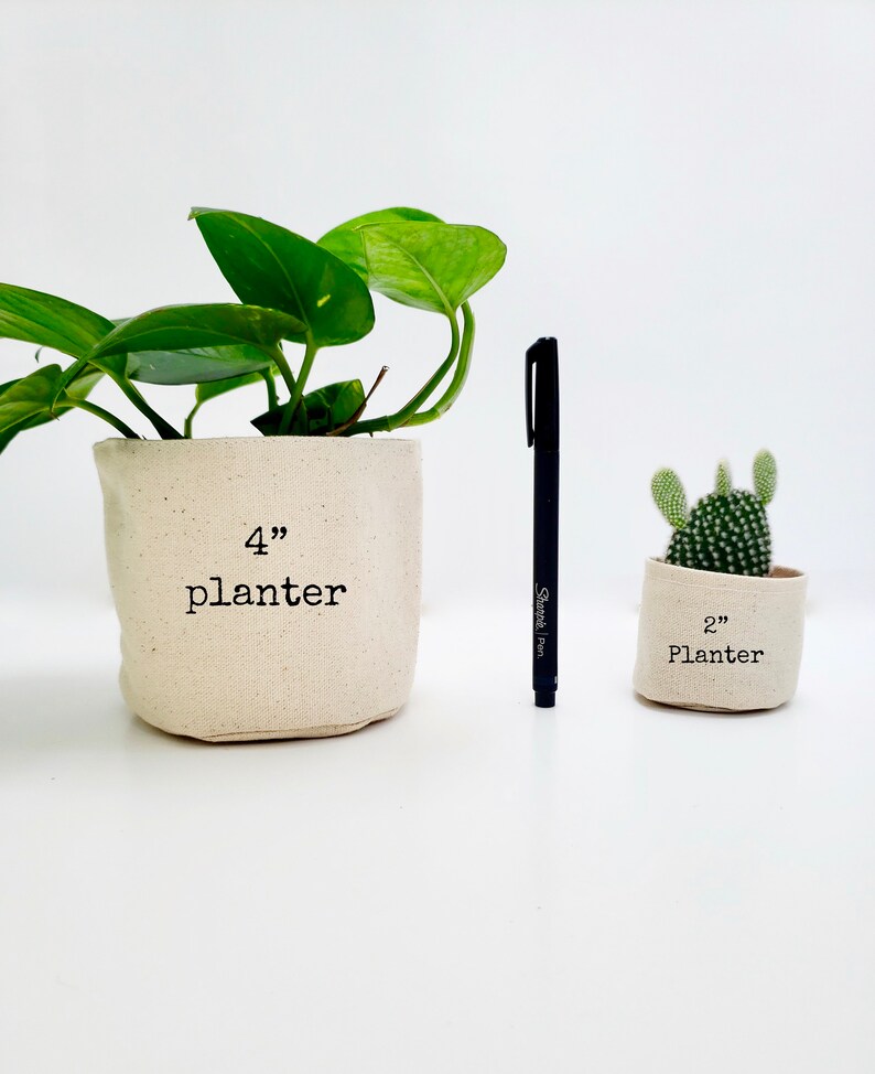 Peut inclure: Deux pots en tissu, l'un de 10 cm de diam&egrave;tre et l'autre de 5 cm de diam&egrave;tre. Le plus grand pot est rempli d'une plante verte, et le plus petit pot est rempli d'un cactus. Les deux pots sont blancs avec du texte noir.