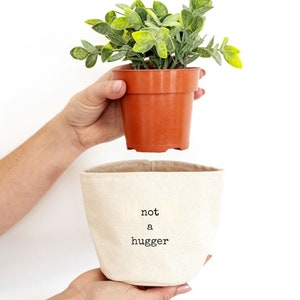 May include: A white canvas planter with the text "not a hugger" in black. A small green plant in a brown plastic pot is being held above the planter.