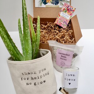 May include: A gift box filled with a potted aloe plant, a bag of lavender bath salts, a small candle, and matches. The plant pot is white canvas with the text "thank you for helping me grow". The candle is clear glass with a white label that says "love, Steve". The bath salts are in a clear plastic bag with a white label that says "Lavender Bath Salts Soak Breath Relax".