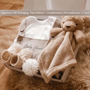 May include: A baby gift set in a white box. The set includes a white onesie, beige overalls with the name "Baby Johnson", knitted booties, a plush teddy bear, and a soft blanket. A white pom-pom is also included. The text "Signature Gift Packaging" is visible.