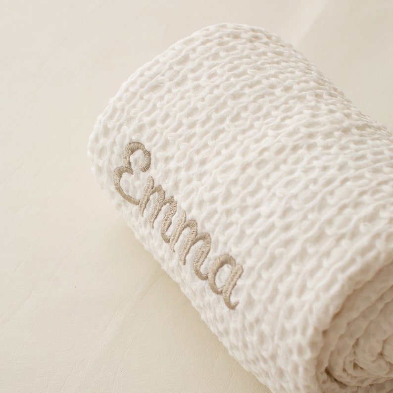 Op de afbeelding: Witte babydeken met een wafelstructuur, met de naam "Emma" geborduurd in beige draad.