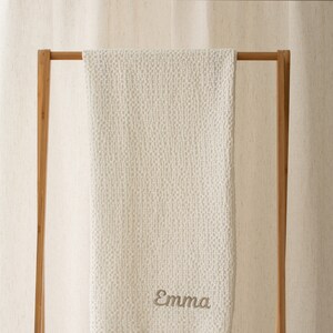 Op de afbeelding: Witte gebreide babydeken met een subtiel textuurpatroon, hangend aan een houten rek. De deken heeft de naam "Emma" geborduurd in lichtbruin garen.