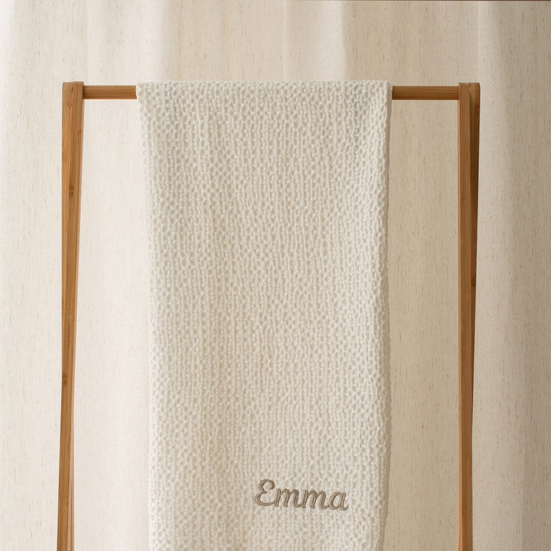 Op de afbeelding: Witte gebreide babydeken met een subtiel textuurpatroon, hangend aan een houten rek. De deken heeft de naam "Emma" geborduurd in lichtbruin garen.