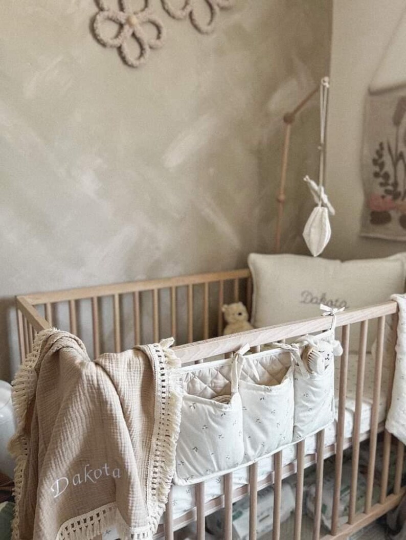 Op de afbeelding: Een houten wieg met een witte en beige gewatteerde wiegorganizer met drie vakken. De organizer heeft een bloemenprint en hangt aan de zijkant van de wieg. Een beige deken met een franje rand en de naam "Dakota" geborduurd erop is over de zijkant van de wieg gelegd.